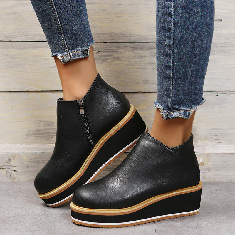 Black platform ankle boots with brown soles worn with blue jeans on a wooden floor.